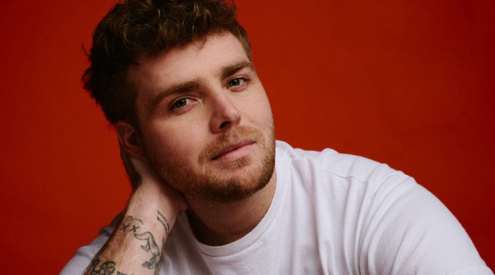 A man in a white tshirt with short brown hair in front of a red background